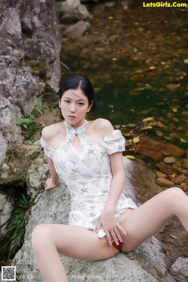 A woman in a white dress sitting on a rock by a stream.